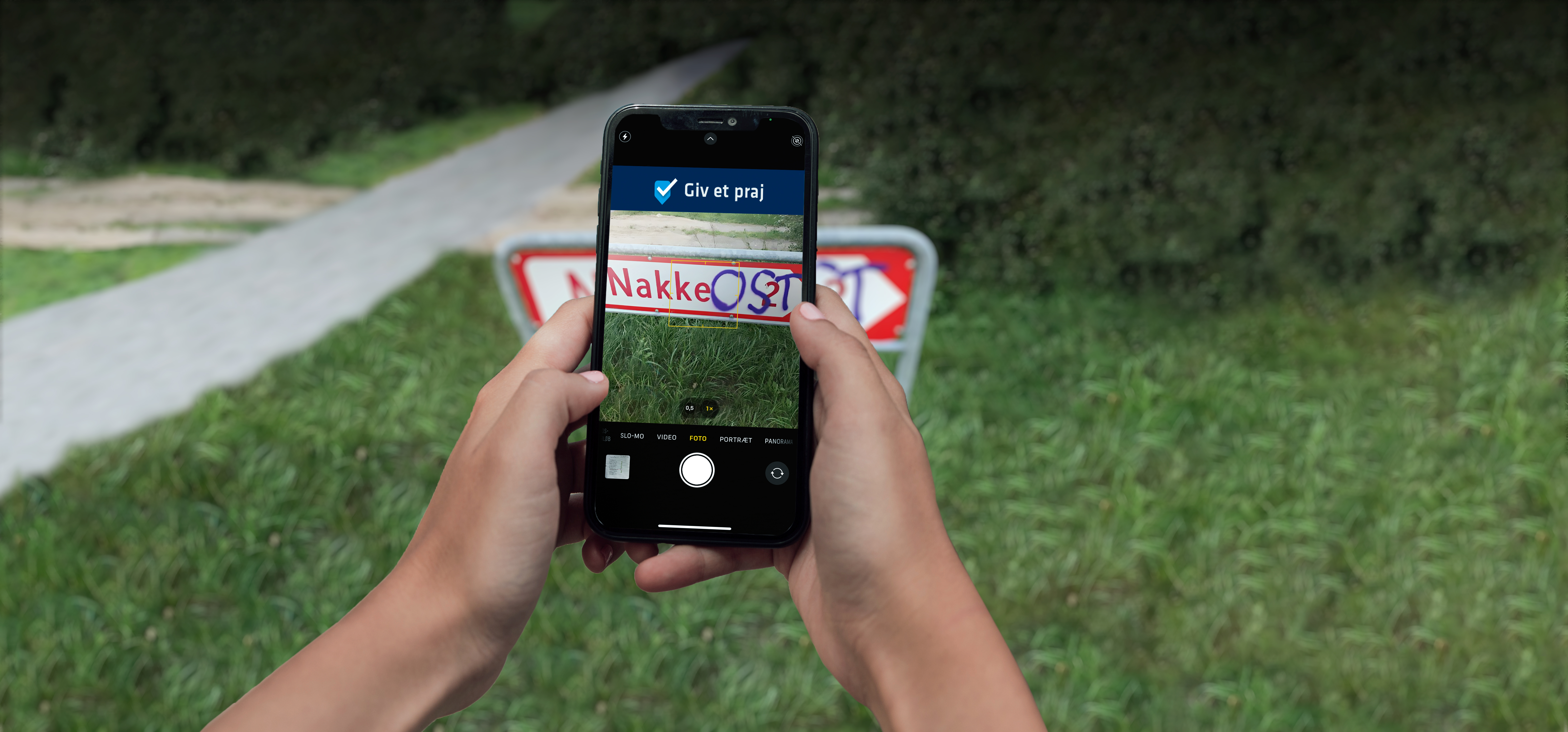 Hænder holder mobiltelefon og tager et billede af et vejskilt hvor der står "Nakke" med henblik på at praje kommunen om graffiti. Ukendte geningsmænd har tilføjet ordet "OST" med graffiti