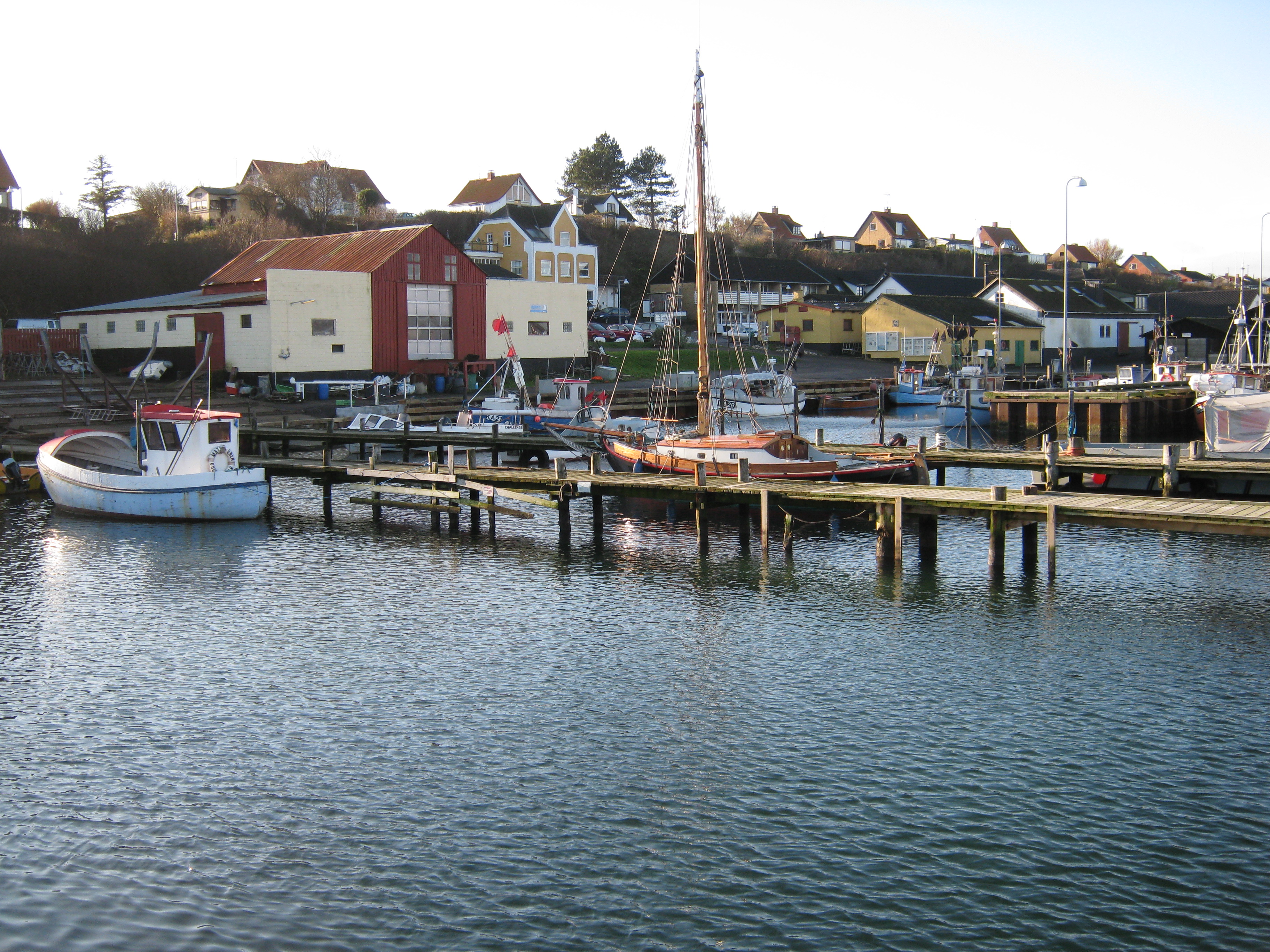 Kig fra vandet mod havnefronten. I forgrunden moler og både. I baggrunden huse på havnen.